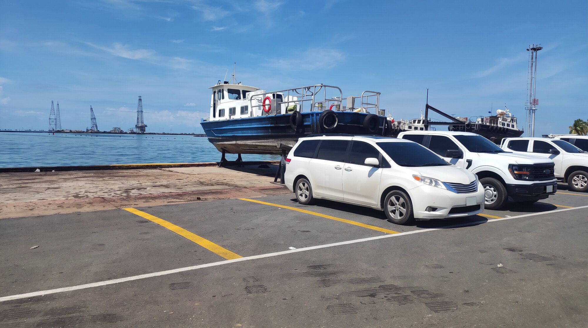 Minivan y camionetas blancas estacionadas en un muelle, con una embarcación en dique y torres petroleras en el lago al fondo